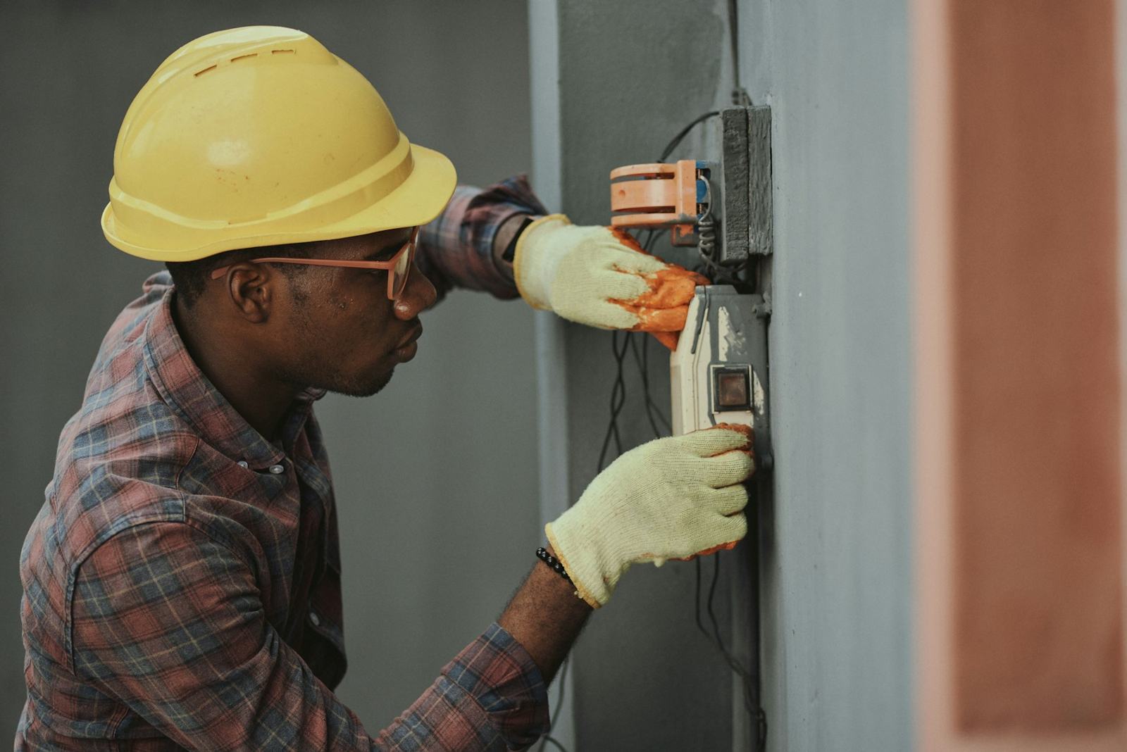 Dan Okafor in a yellow hard hat, safety glasses and work gloves testing a wall-mounted electricity meter