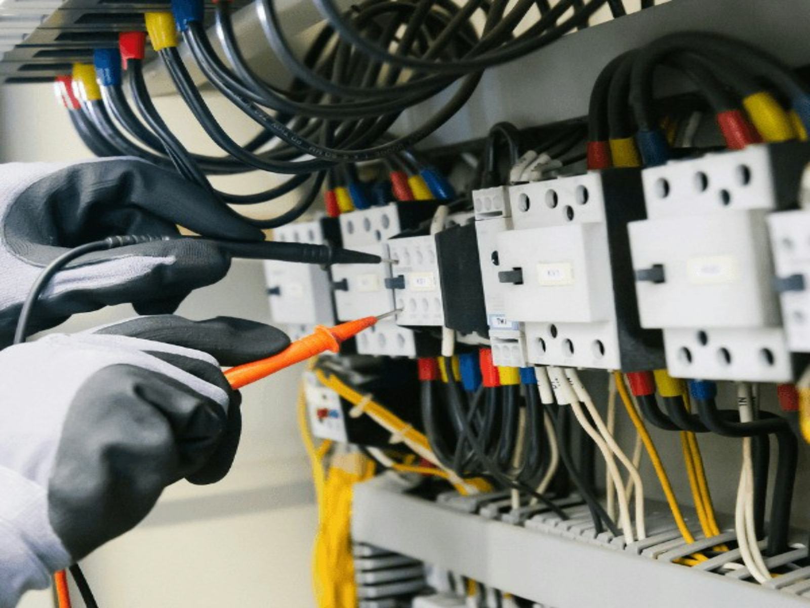 Gloved hands holding red and black test probes against circuit breakers in a switchboard full of colour-coded wiring