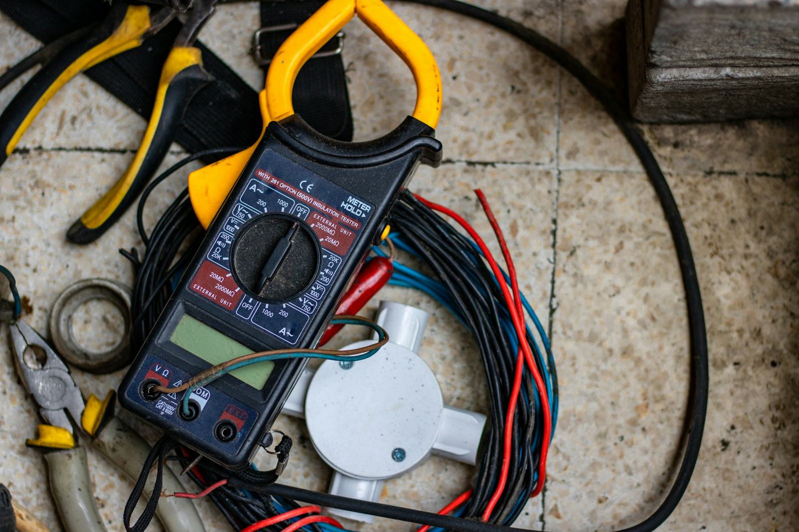 Clamp meter, pliers, a junction box and coils of red, blue and black cable laid out on a tiled floor