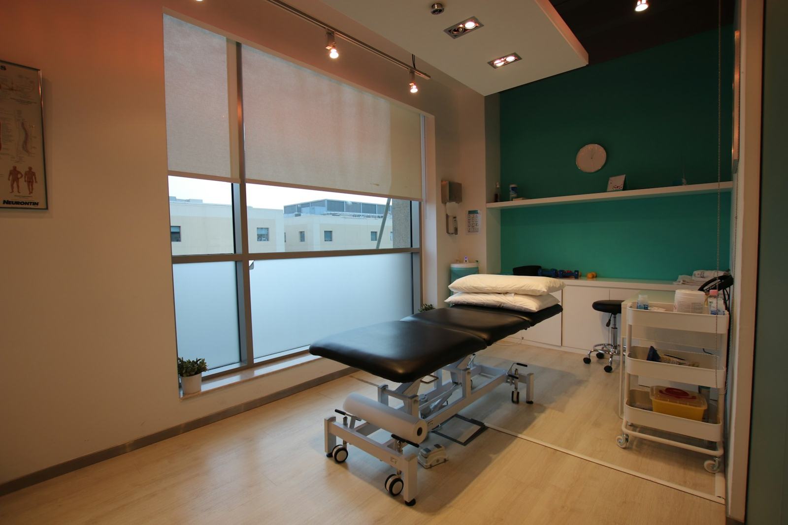 A private treatment room with a black treatment table, a green feature wall and a large window with soft blinds