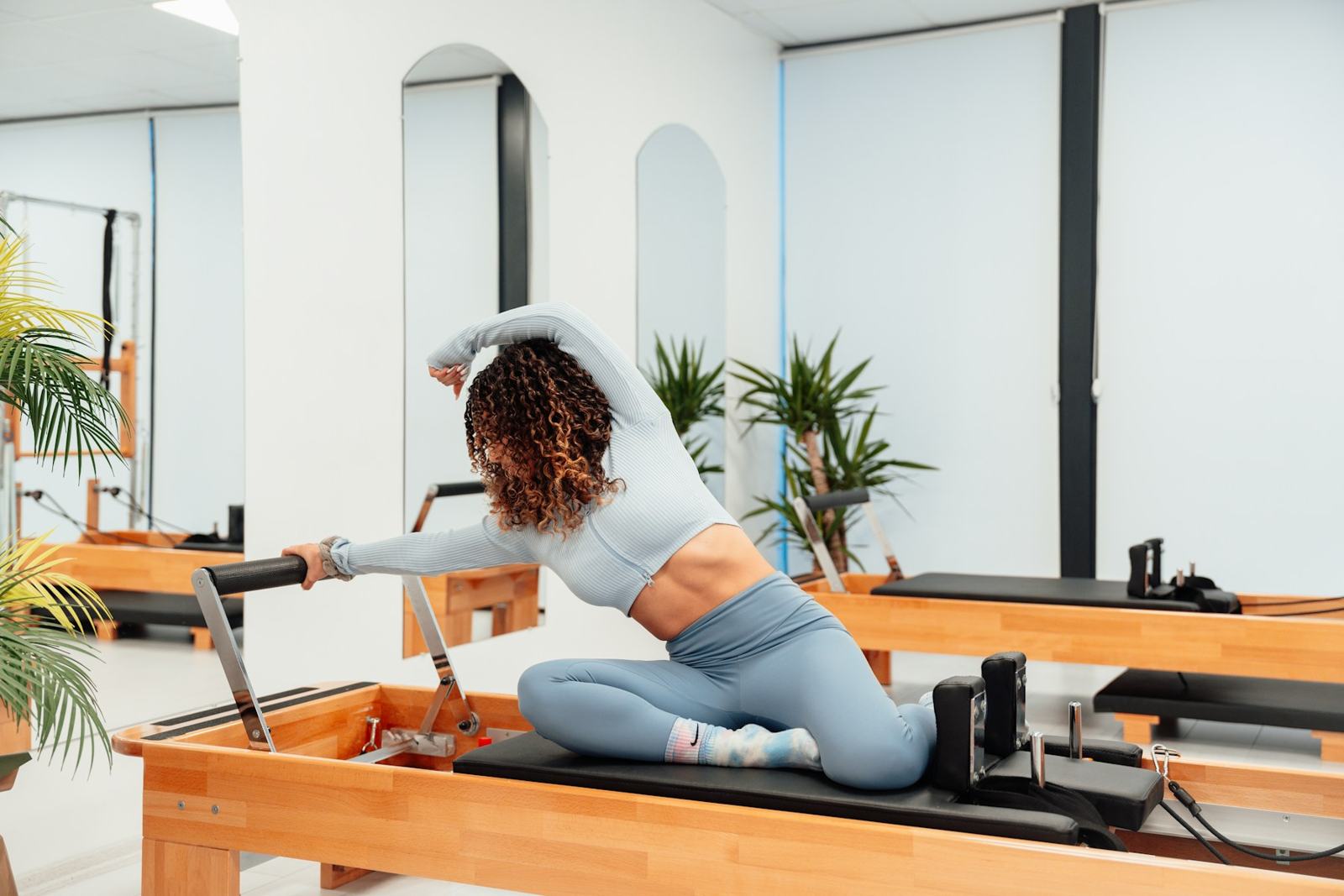 A member in a soft grey set leans into a side stretch on a timber reformer in a bright white studio, palms by the window